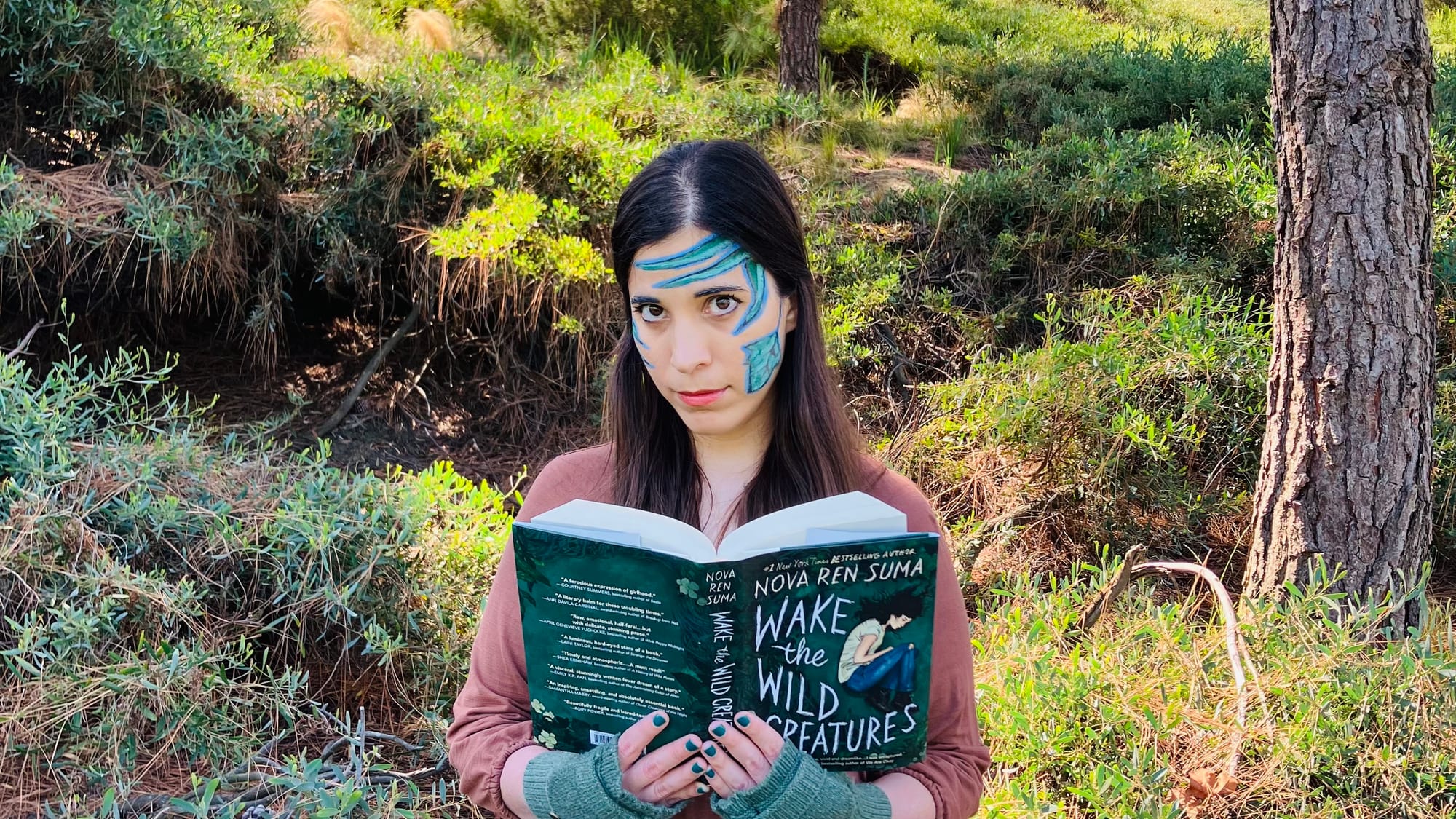 A-M holding Nova Ren Suma’s novel WAKE THE WILD CREATURES. They are standing in front of trees and undergrowth and have blue and green vines painted on their face. They are looking at the camera with the book open.