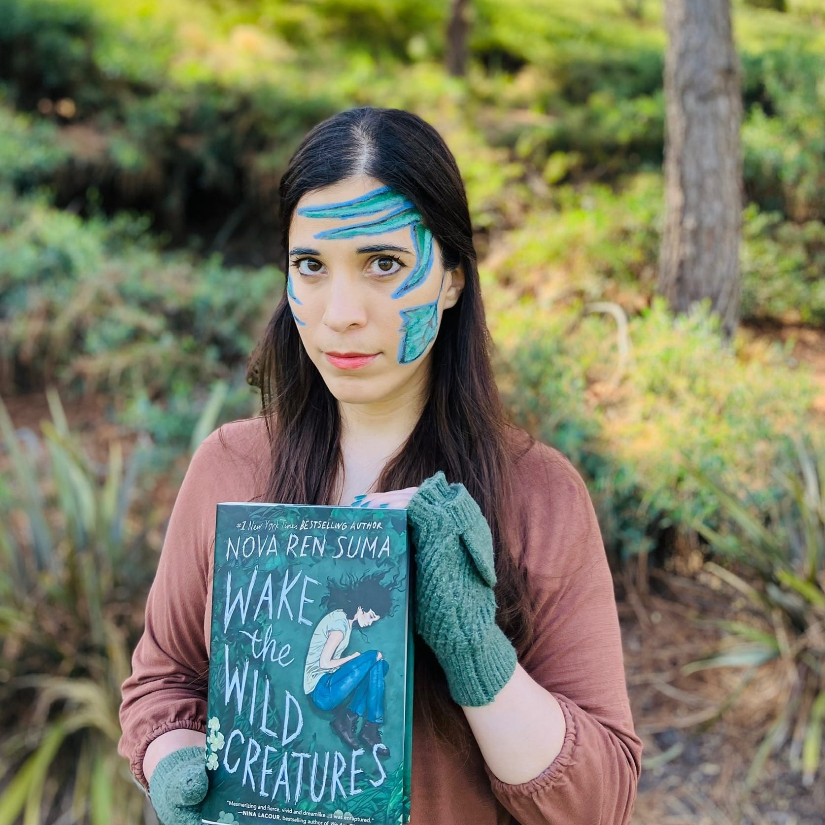 A-M holding the cover of Nova Ren Suma’s novel WAKE THE WILD CREATURES up to the camera. They are standing in front of trees and undergrowth and have blue and green vines painted on their face.
