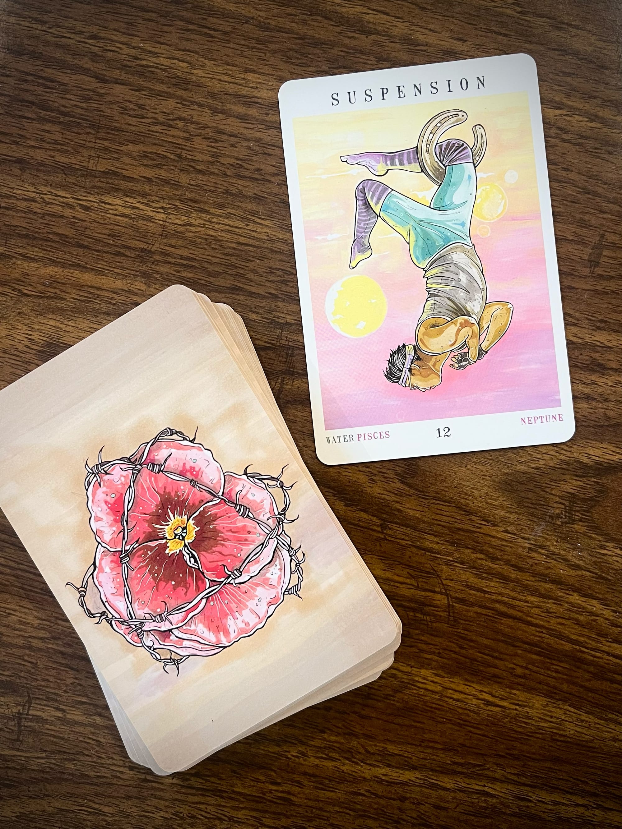 Stack of tarot cards (from The Next World Tarot by Cristy Road) and the specific card that was drawn: number 12, "Suspension," featuring an illustration of a person hanging upside down like an aerial artist with their right knee hooked through a horseshoe, their arms folded into their chest, and a golden sphere behind them that might be a sun