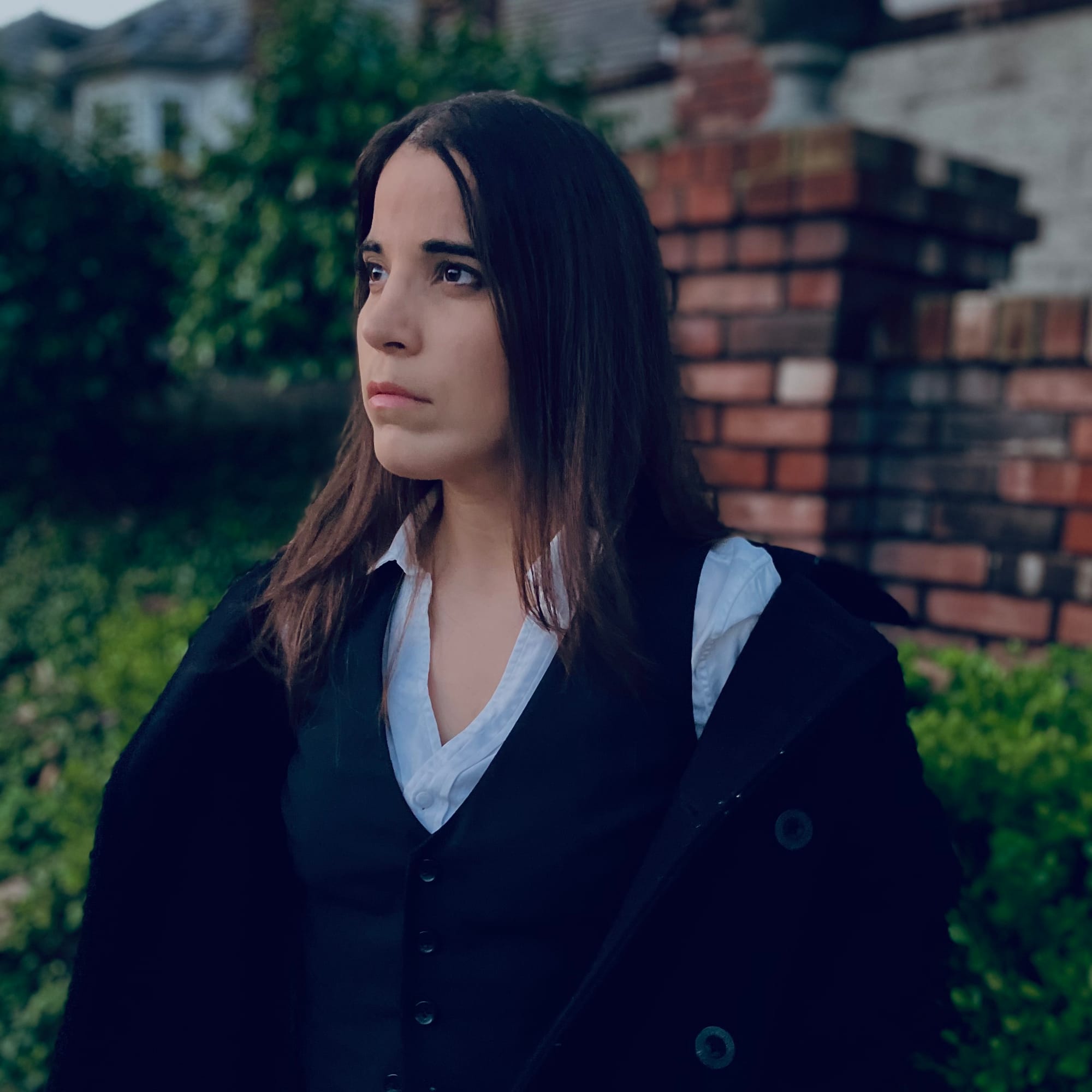 A-M wearing a white collared shirt, black vest, and black coat, standing in front of a brick pillar and hedges on an overcast day. 