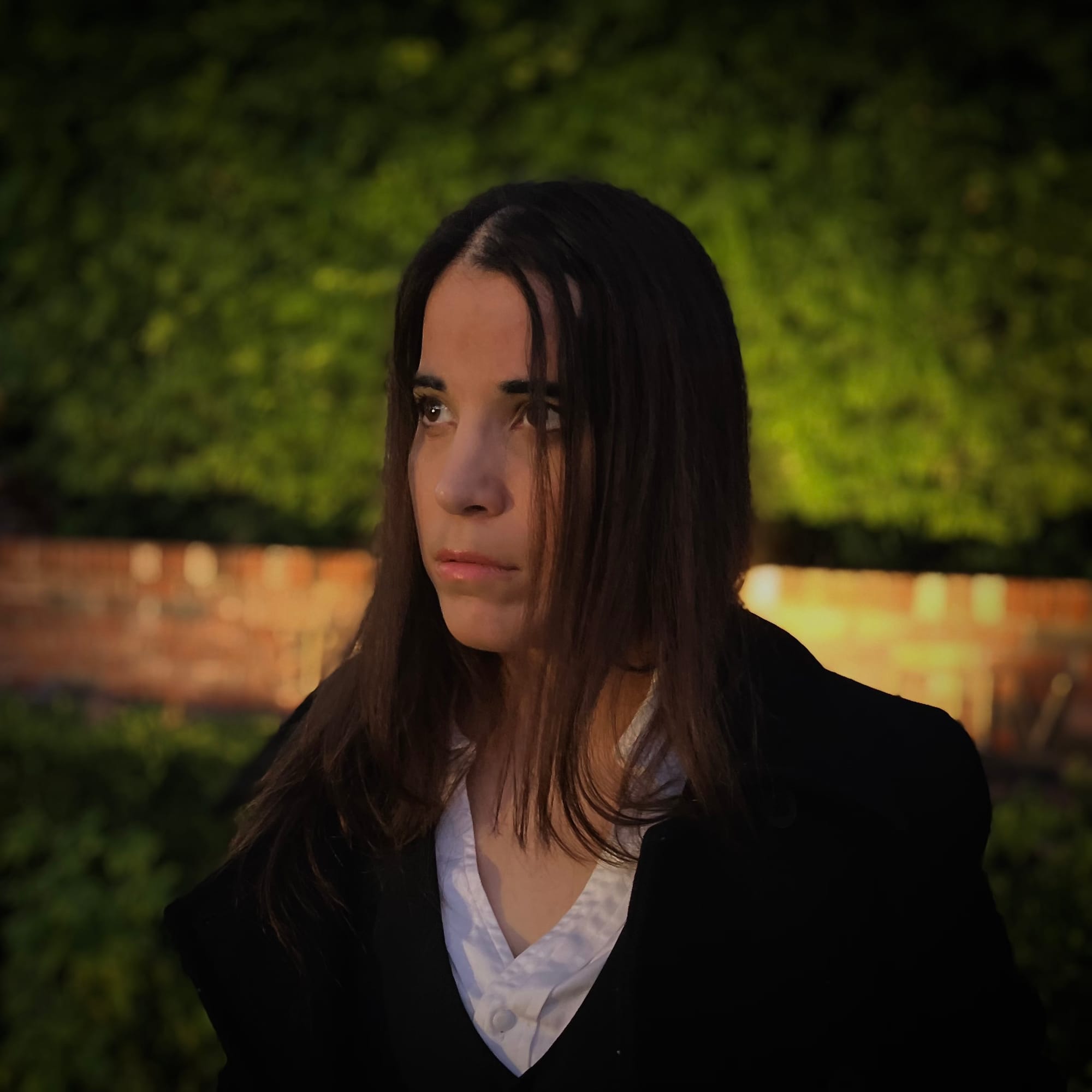 A-M wearing a white collared shirt, black vest, and black coat, standing in front of a brick pillar and hedges at night. 