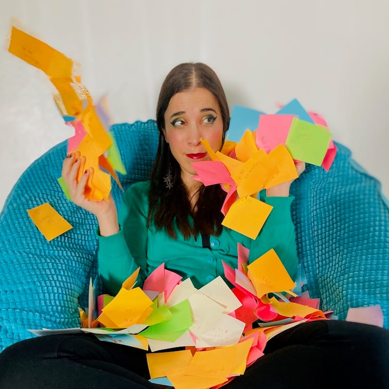 A-M sitting in a teal chair, wearing green eyeshadow and a green sweater, with a flurry of neon sticky notes around them.