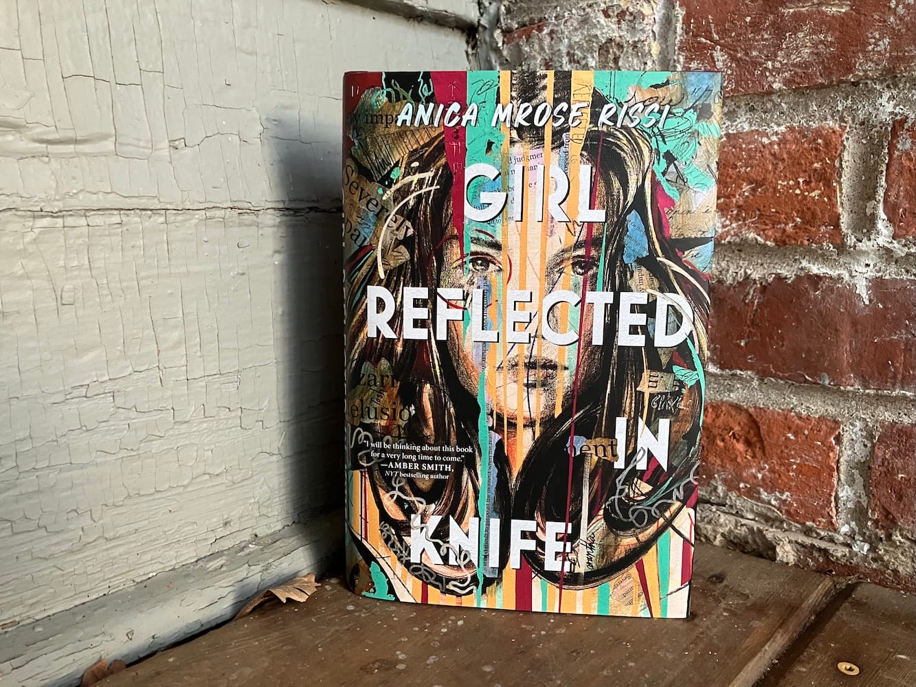 A hardcover of Girl Reflected in Knife on a wooden floor, near the corner where a brick wall and a painted wall meet.